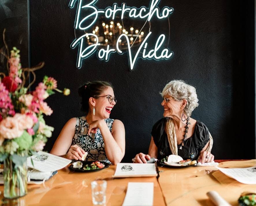 Two guests laughing at Borracho's, under the Borracho Por Vida neon sign